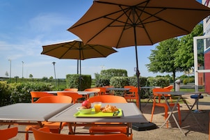 An outdoor seating area with orange chairs and tables, umbrellas, and a table with a croissant, fruit, and juice.