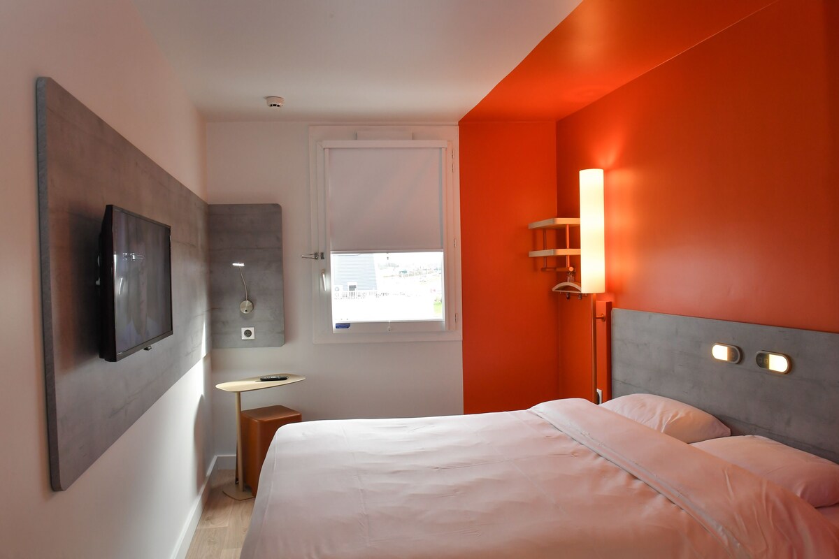 Una habitación de hotel moderna con una cama grande, un televisor montado en la pared, una mesita y un ventanal con vistas al exterior.