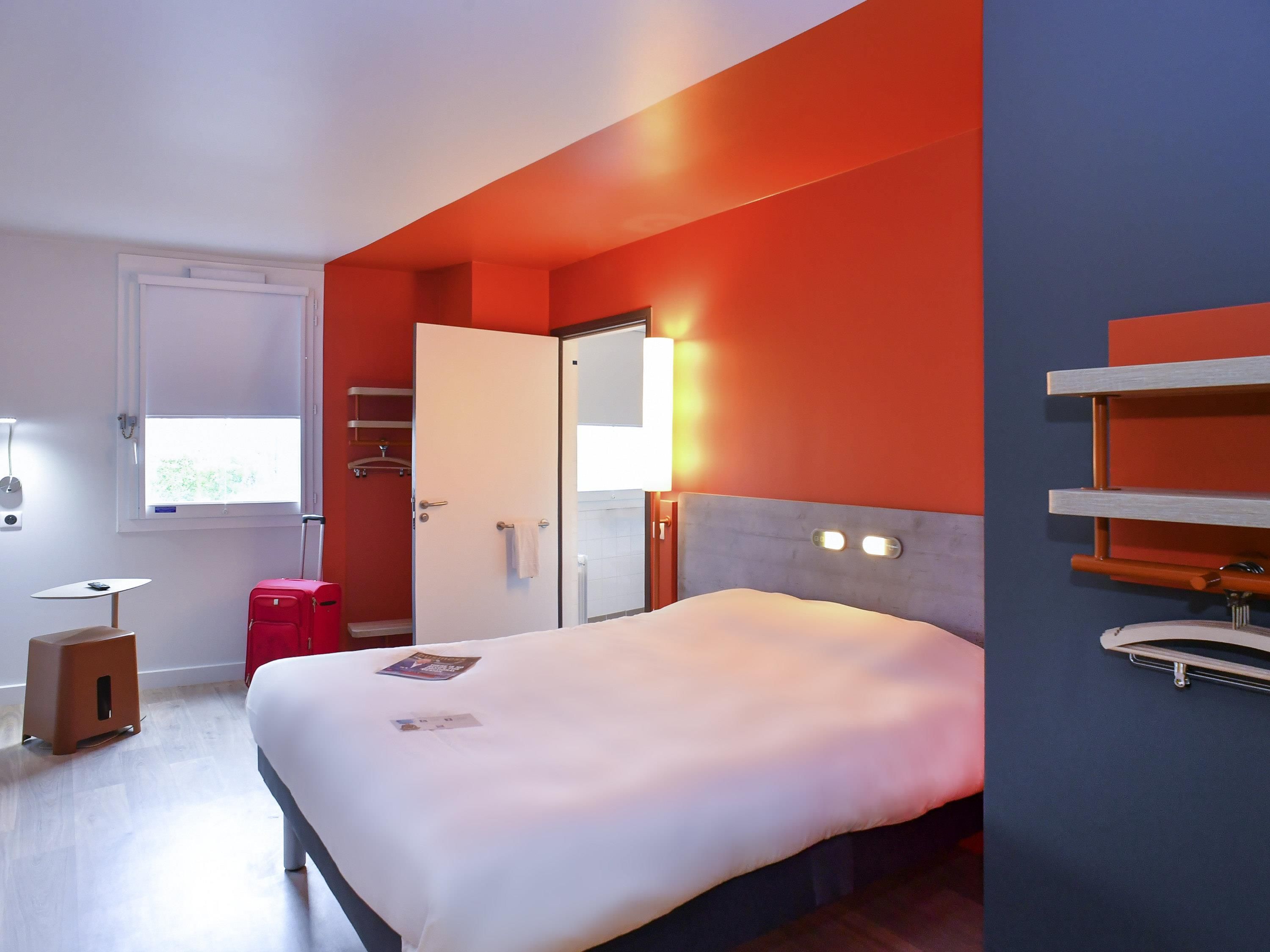 Une chambre d’hôtel moderne avec un lit, une table de chevet, un petit tabouret, une valise rouge, une fenêtre donnant sur un paysage agréable et une porte de salle de bain.