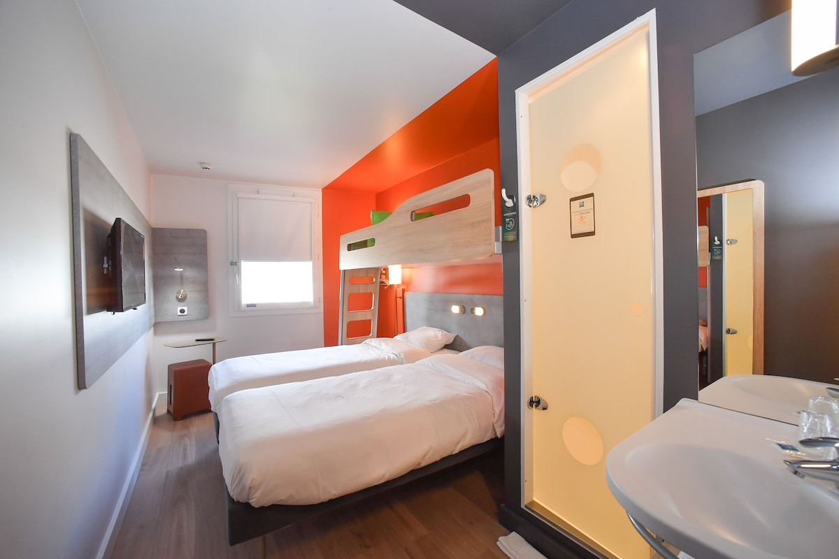 Una habitación de hotel con dos camas, una litera, un baño con lavamanos y una pequeña mesa con una silla.