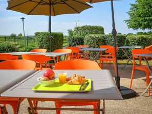 Un área de comedor al aire libre con mesas y sillas, una mesa con alimentos para el desayuno, y un paraguas grande.