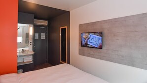 Habitación de hotel con cama, televisor montado en la pared, baño con lavamanos y espejo, y unidad de aire acondicionado.
