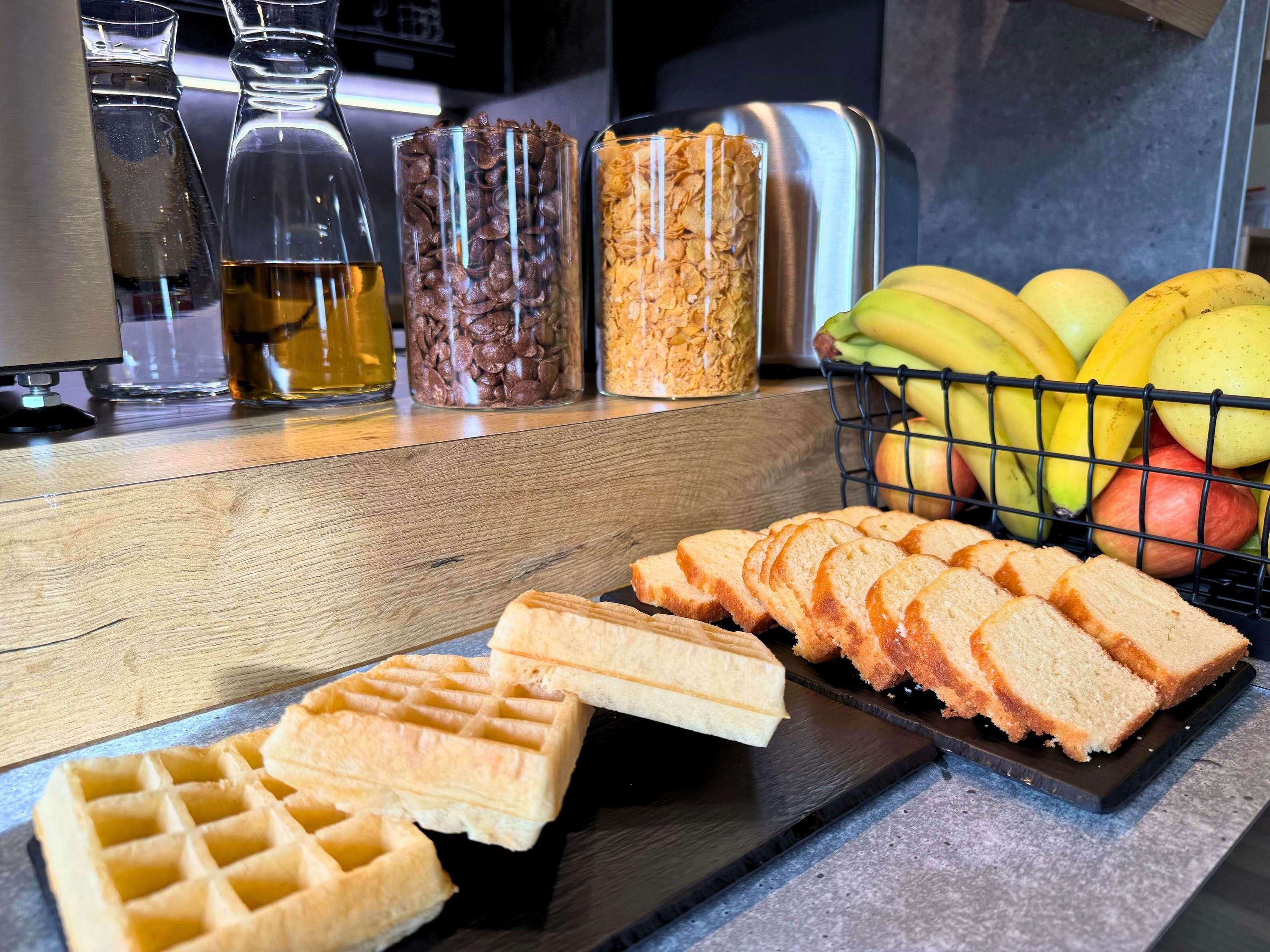 A kitchen counter with waffles, sliced bread, a fruit basket, and jars of condiments.