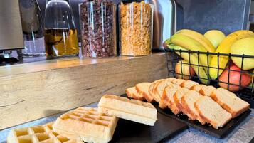 A kitchen counter with waffles, sliced bread, a fruit basket, and jars of condiments.