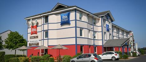 Un hotel de dos pisos con un letrero que dice ibis budget, indicando que es un hotel económico. Hay sombrillas y un estacionamiento con coches.