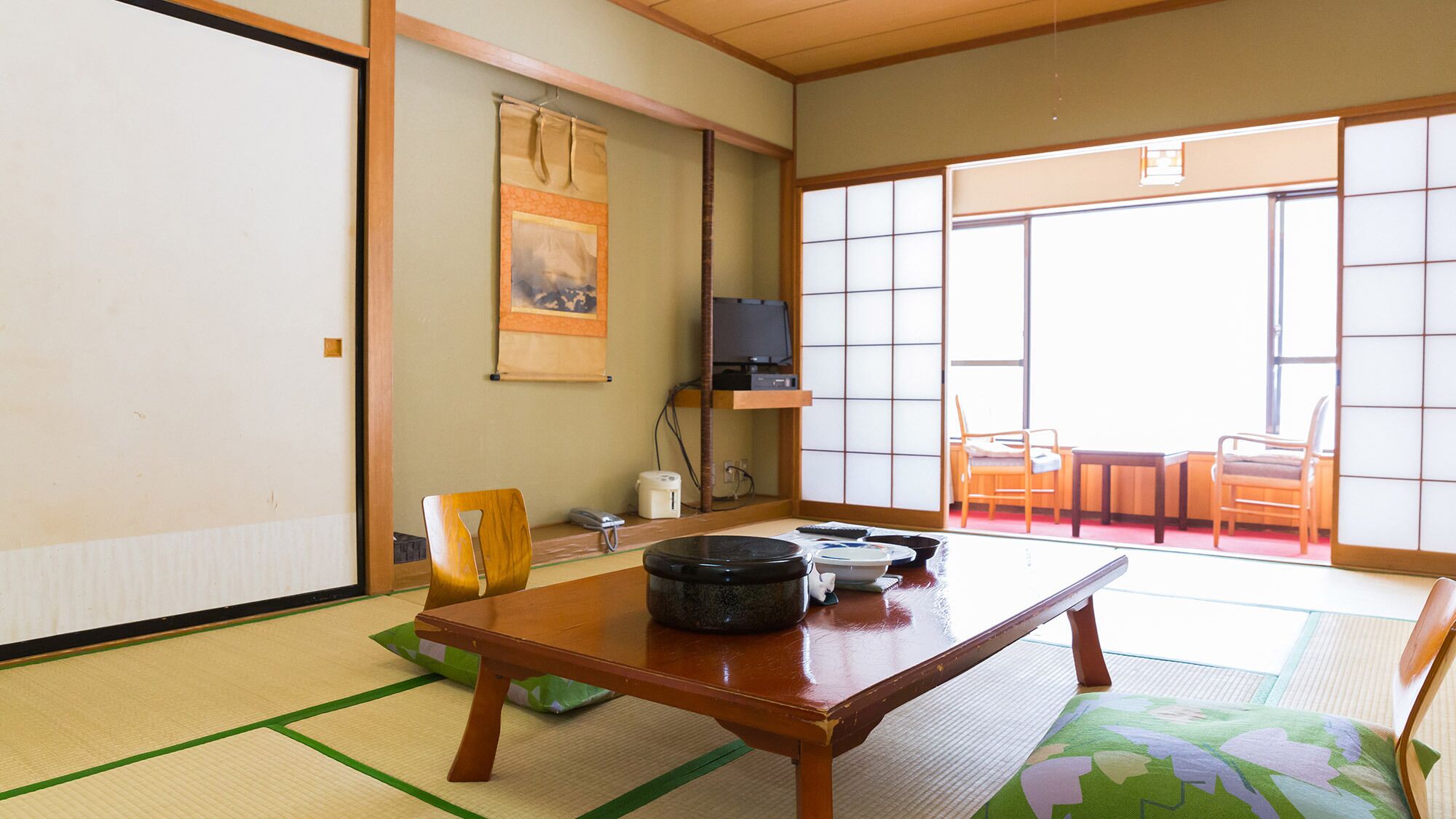 A traditional Japanese room with tatami flooring, a low table, chairs, a television, and a sliding door leading to an outdoor area.