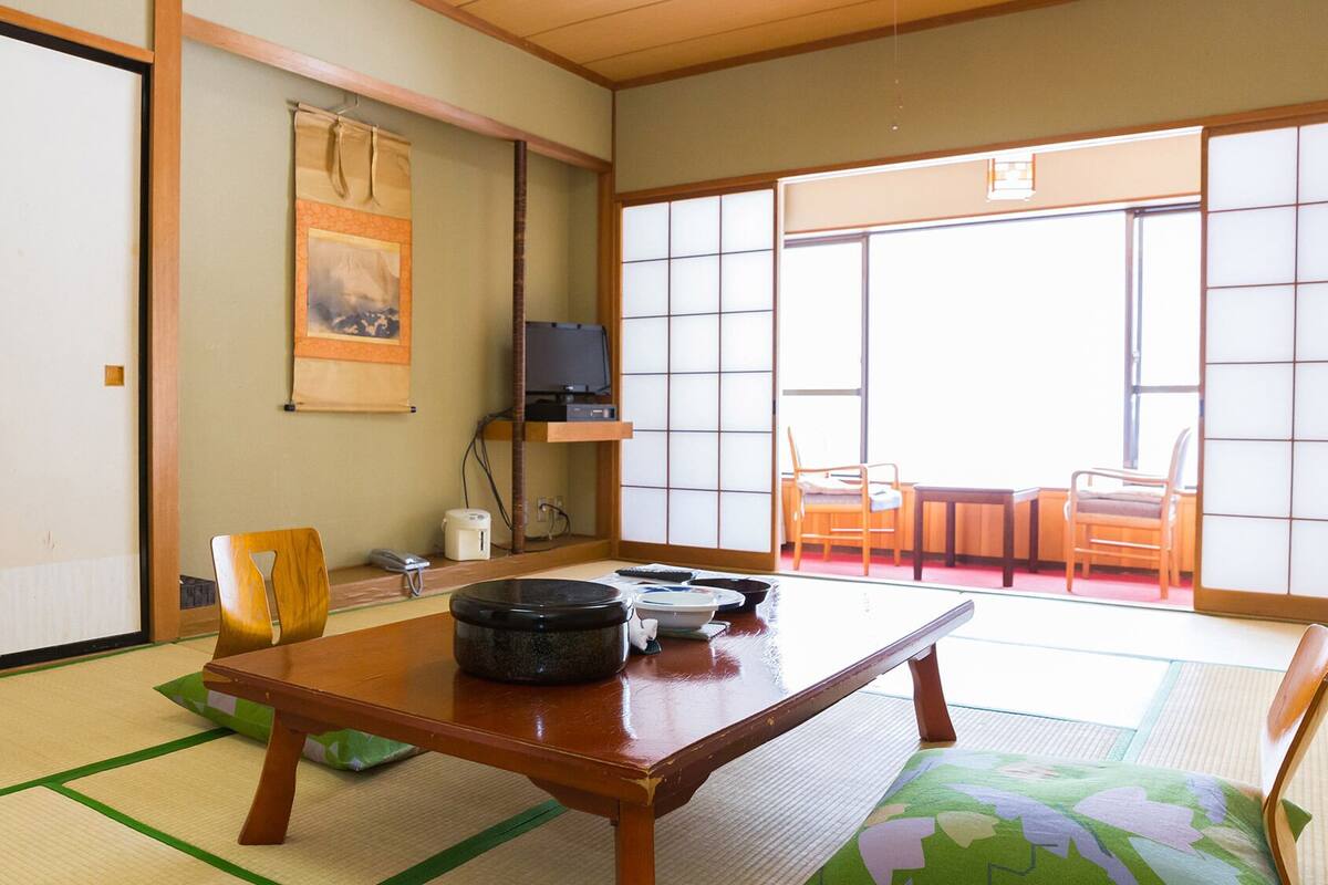 Ein traditioneller japanischer Raum mit Tatami-Bodenbelag, einem niedrigen Tisch, Stühlen, einem Fernseher und einer Schiebetür zu einem Außenbereich.
