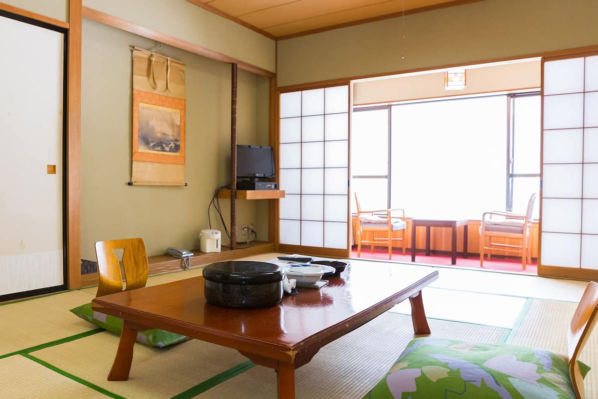 A traditional Japanese room with tatami flooring, a low table, chairs, a television, and a sliding door leading to an outdoor area.