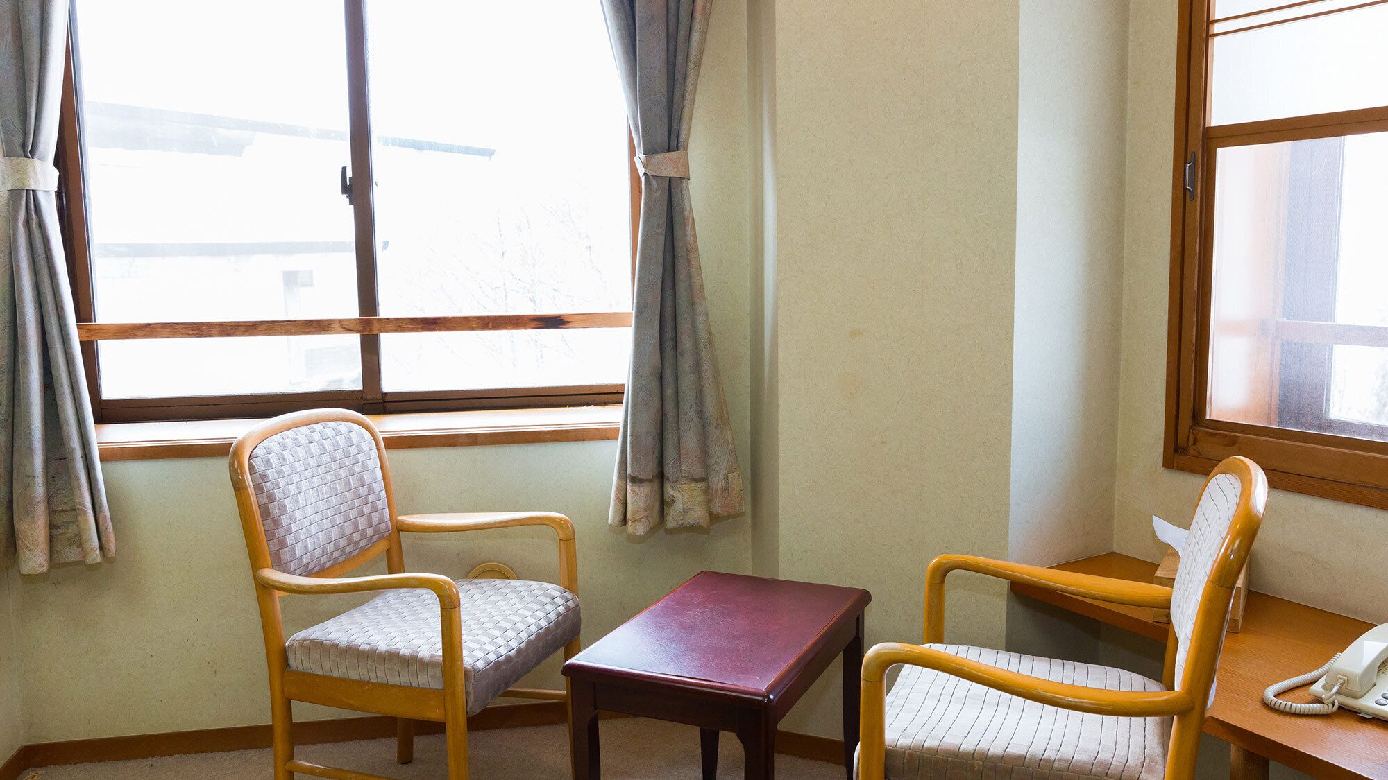 A room with two wooden chairs, a small table, a telephone, and a window with curtains.