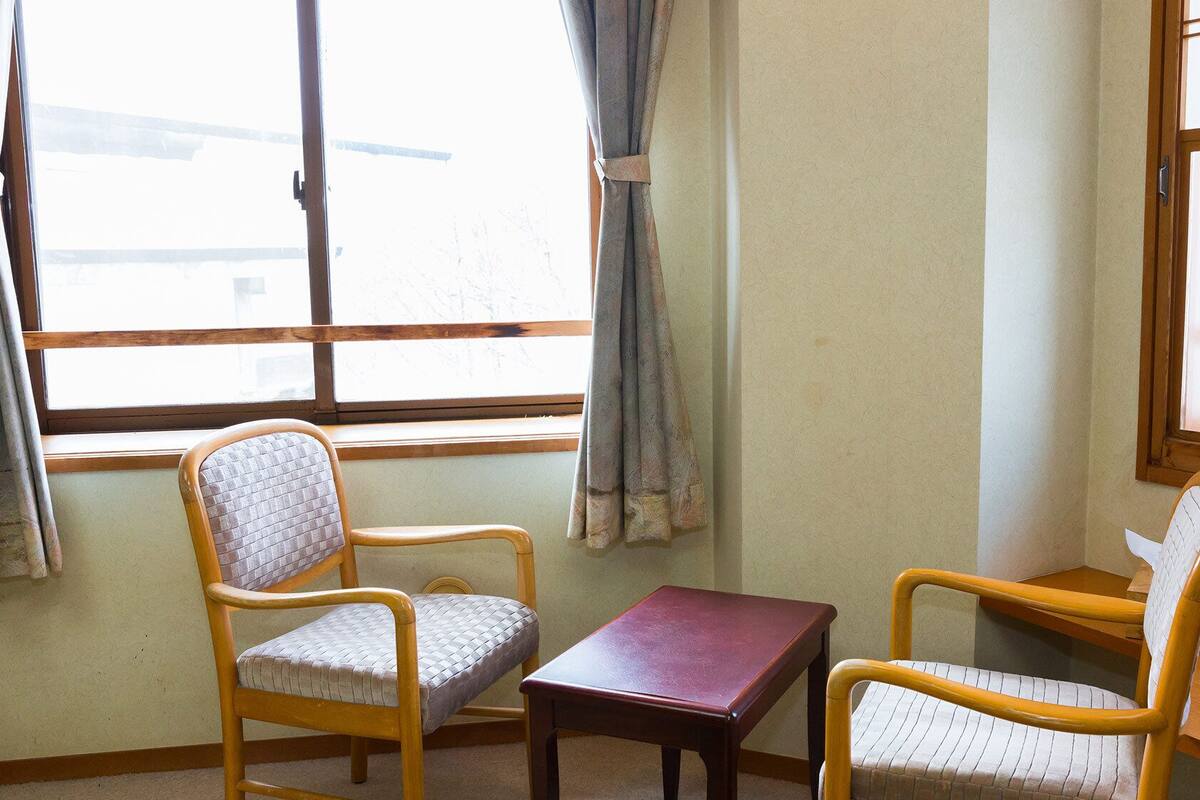 Ein Zimmer mit zwei Holzstühlen, einem kleinen Tisch, einem Telefon und einem Fenster mit Vorhängen.