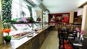 Ein Bereich mit Buffet und vielfältigen Speisen, eine Sitzecke mit Tischen und Stühlen sowie dekorative Pflanzen.