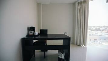 A small, dark-colored table with a chair, a coffee maker, and a trash bin in a room with a view of a cityscape through a window with curtains.