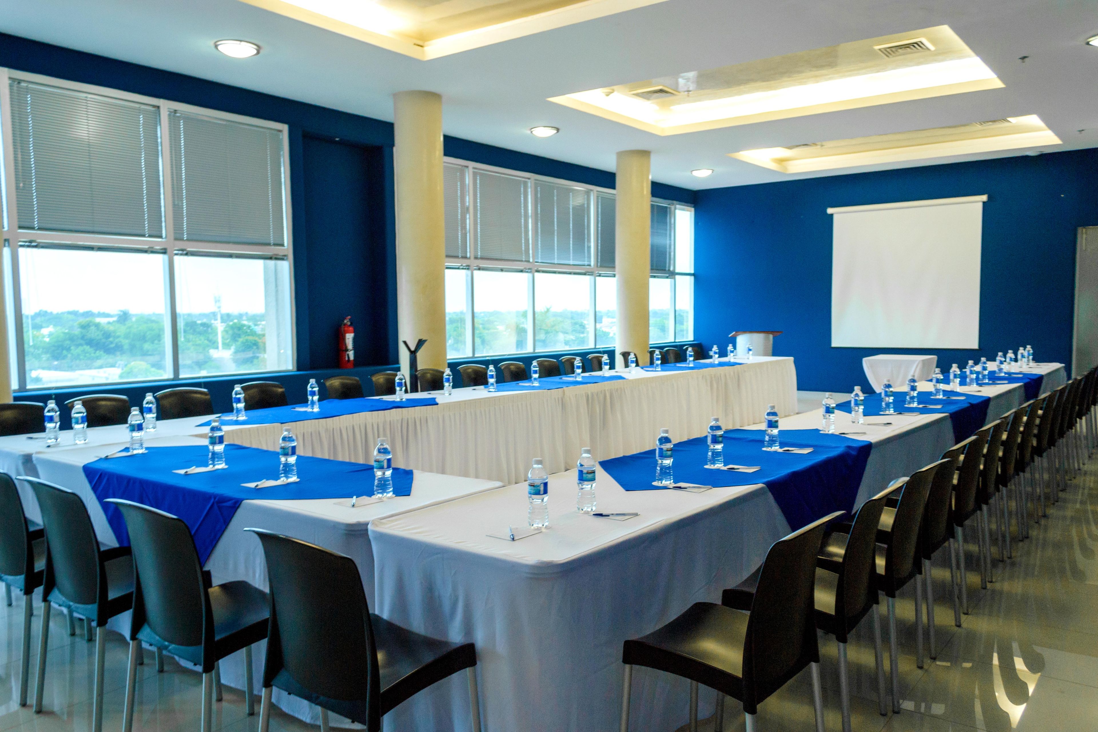 Una sala conferenze con lunghe tavolate, sedie, bottiglie d'acqua e uno schermo per proiezioni.