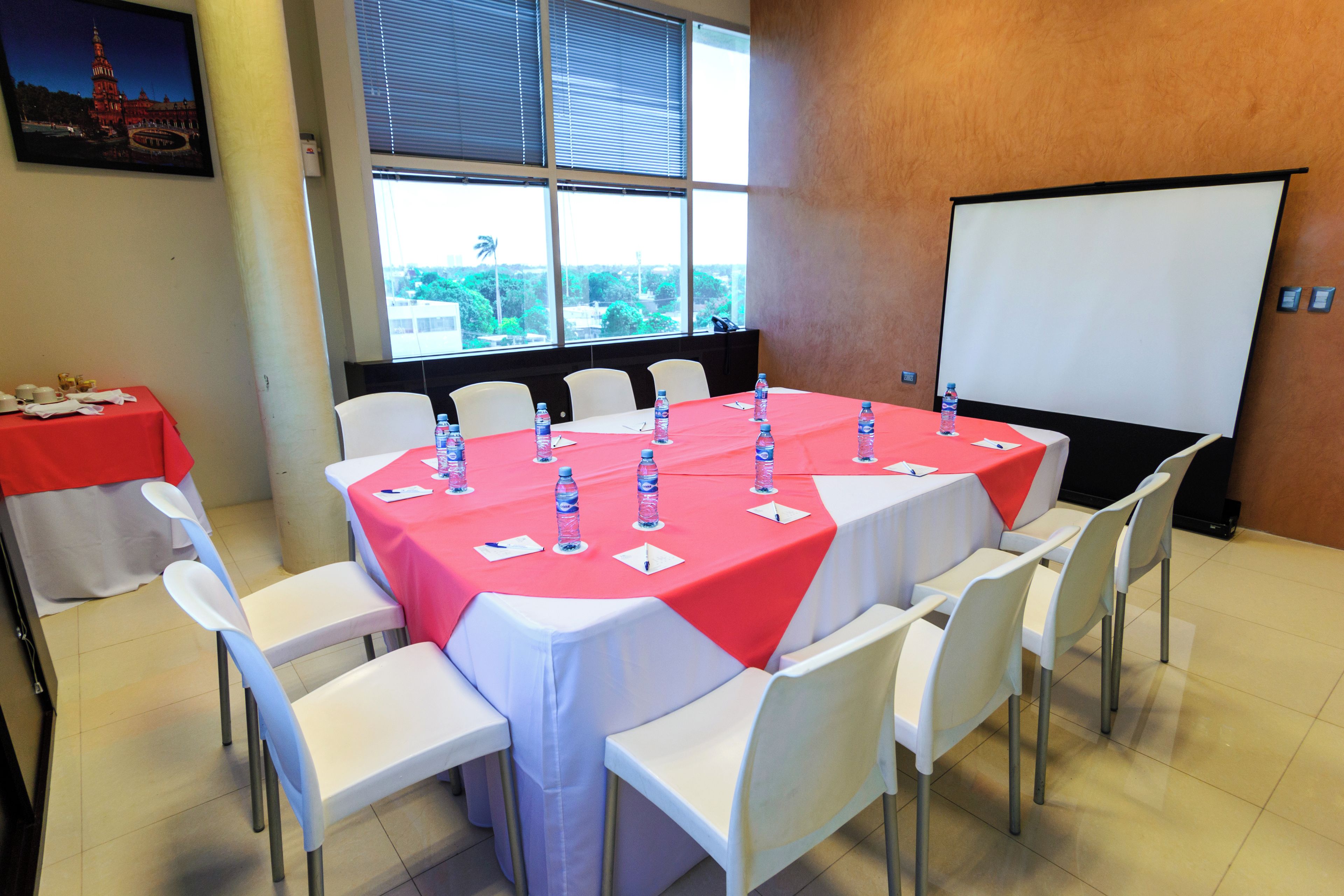 A conference room with a large table covered in a red tablecloth, surrounded by white chairs, and a projector screen on the wall.