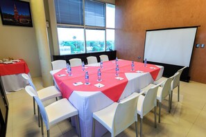 Uma sala de conferências com uma mesa grande coberta por toalha vermelha, rodeada por cadeiras brancas, e uma tela de projetor na parede.