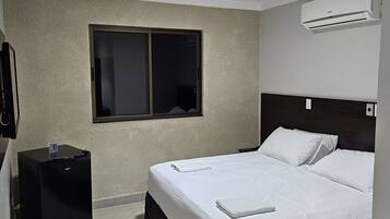 Una habitación de hotel con cama, una mesita de noche negra pequeña, una ventana y un aire acondicionado.