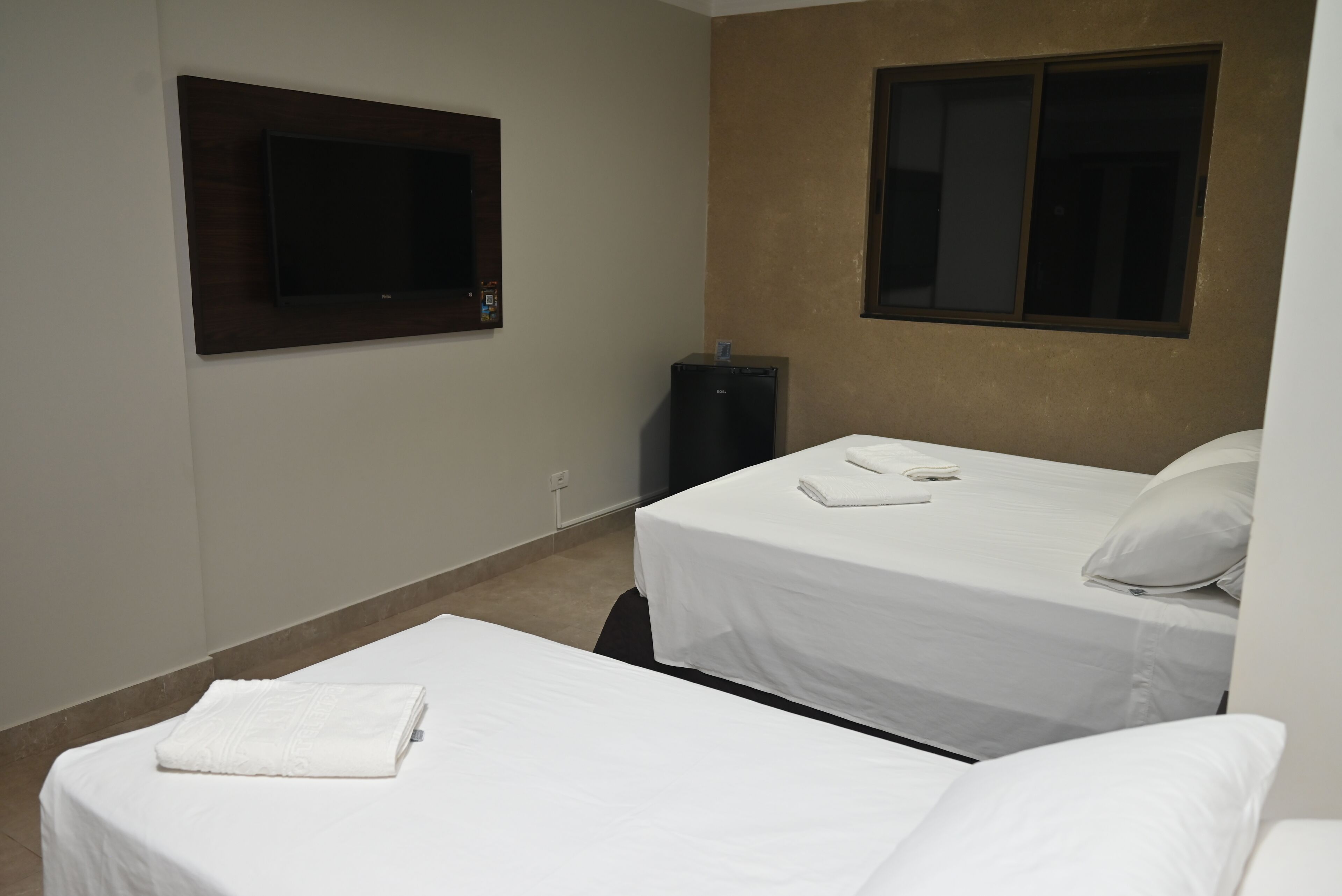 A hotel room with two single beds, a flat-screen TV mounted on the wall, a small refrigerator, and a window.