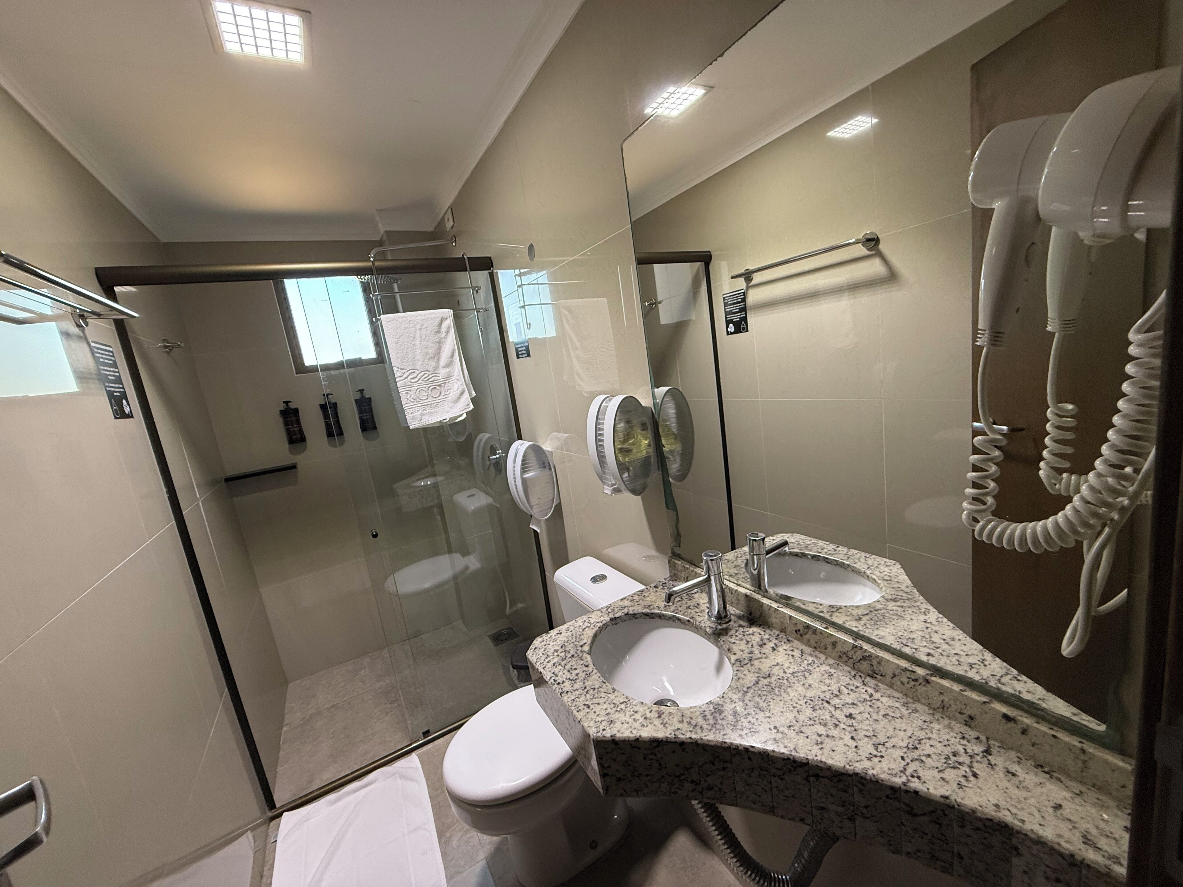 A modern bathroom with a glass-enclosed shower, a white toilet, a granite countertop with a sink, and a hairdryer mounted on the wall.