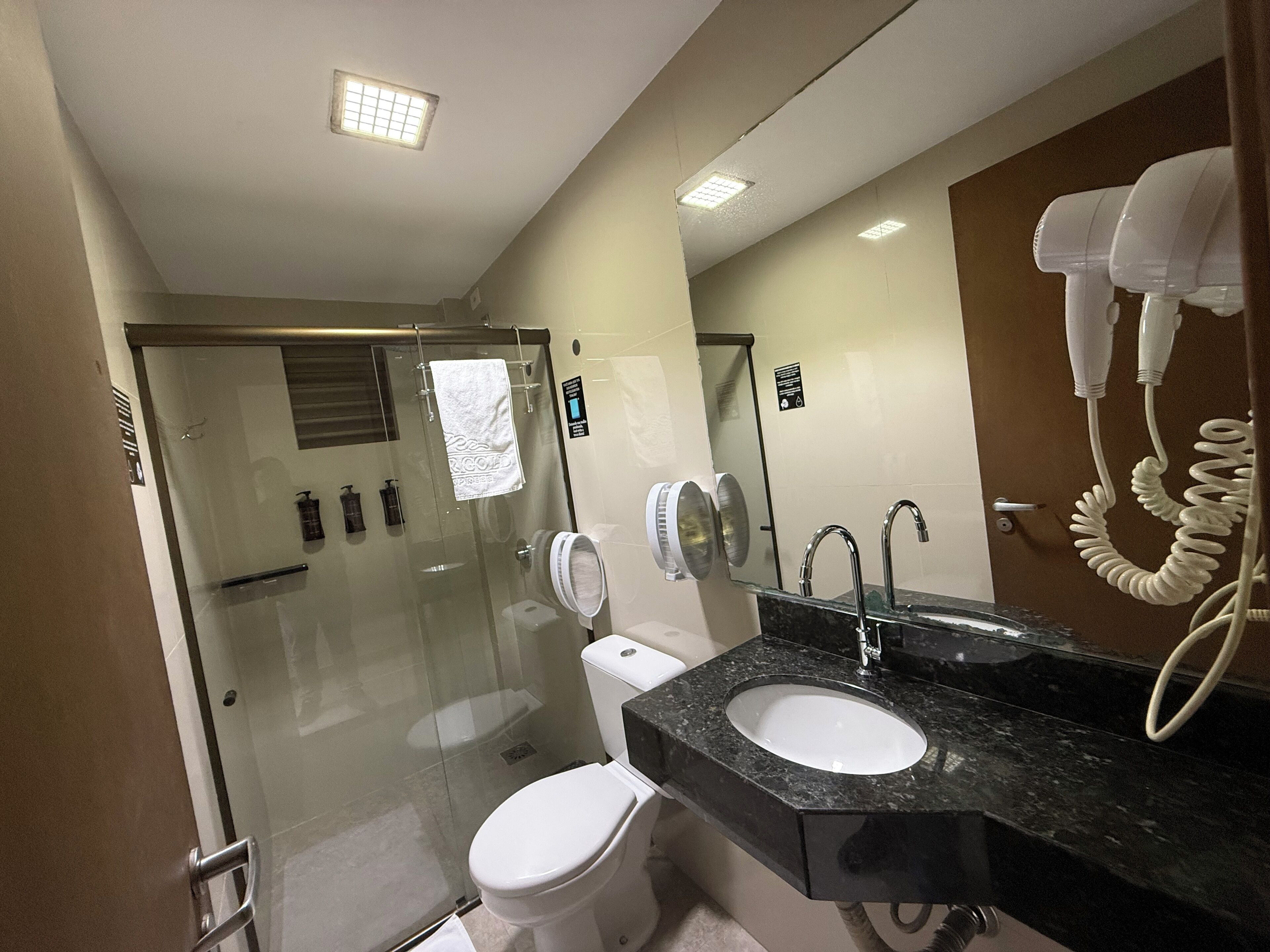 Un baño moderno con cabina de ducha de vidrio, un inodoro blanco, un lavabo con encimera negra y un secador de pelo montado en la pared.
