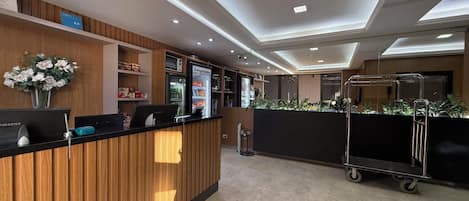 A modern reception area with a wooden reception desk, a Samsung monitor, a refrigerator, and a cart with a plant.