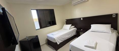 A hotel room with two single beds, a desk, a chair, a TV, and an air conditioning unit.