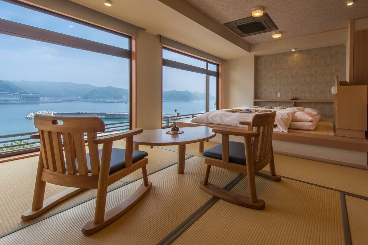 A room with a bed, a table, and chairs, large windows with a view of a body of water and mountains, and wooden cabinets.