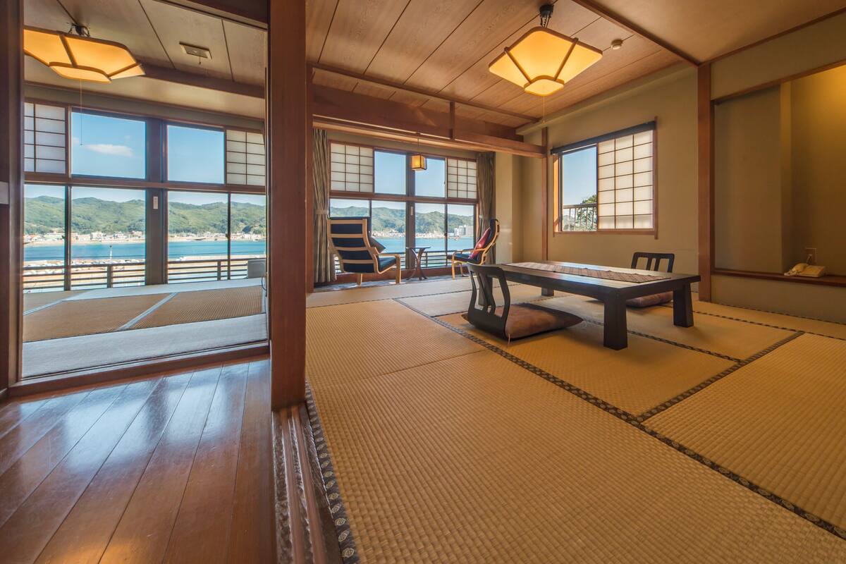 A tatami mat room with a view of the sea, a table, chairs, and a television.