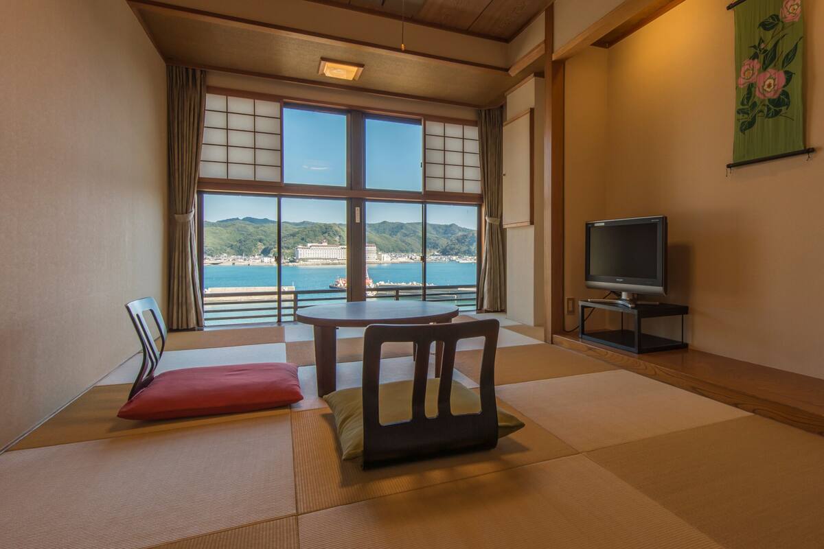A room with tatami flooring, a low table, a TV stand, and a view of a coastal area.