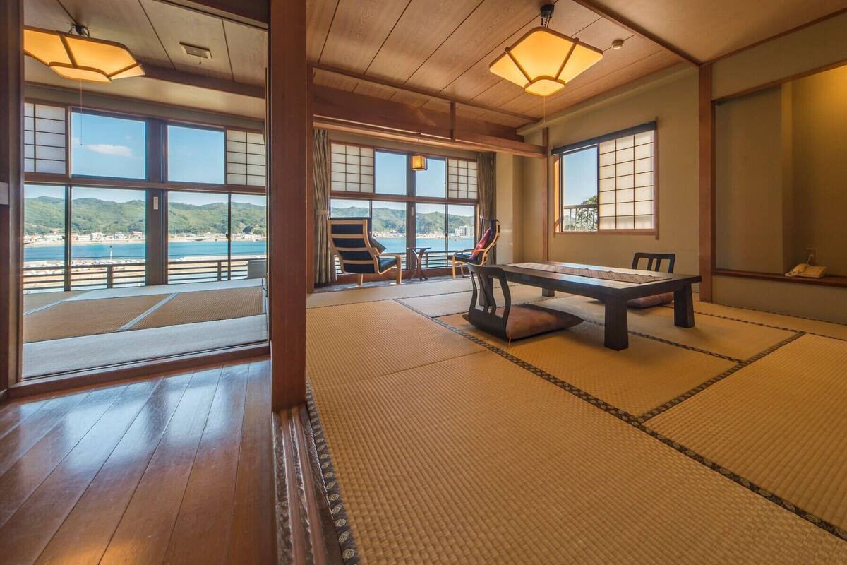A tatami mat room with a view of the sea, a table, chairs, and a TV.