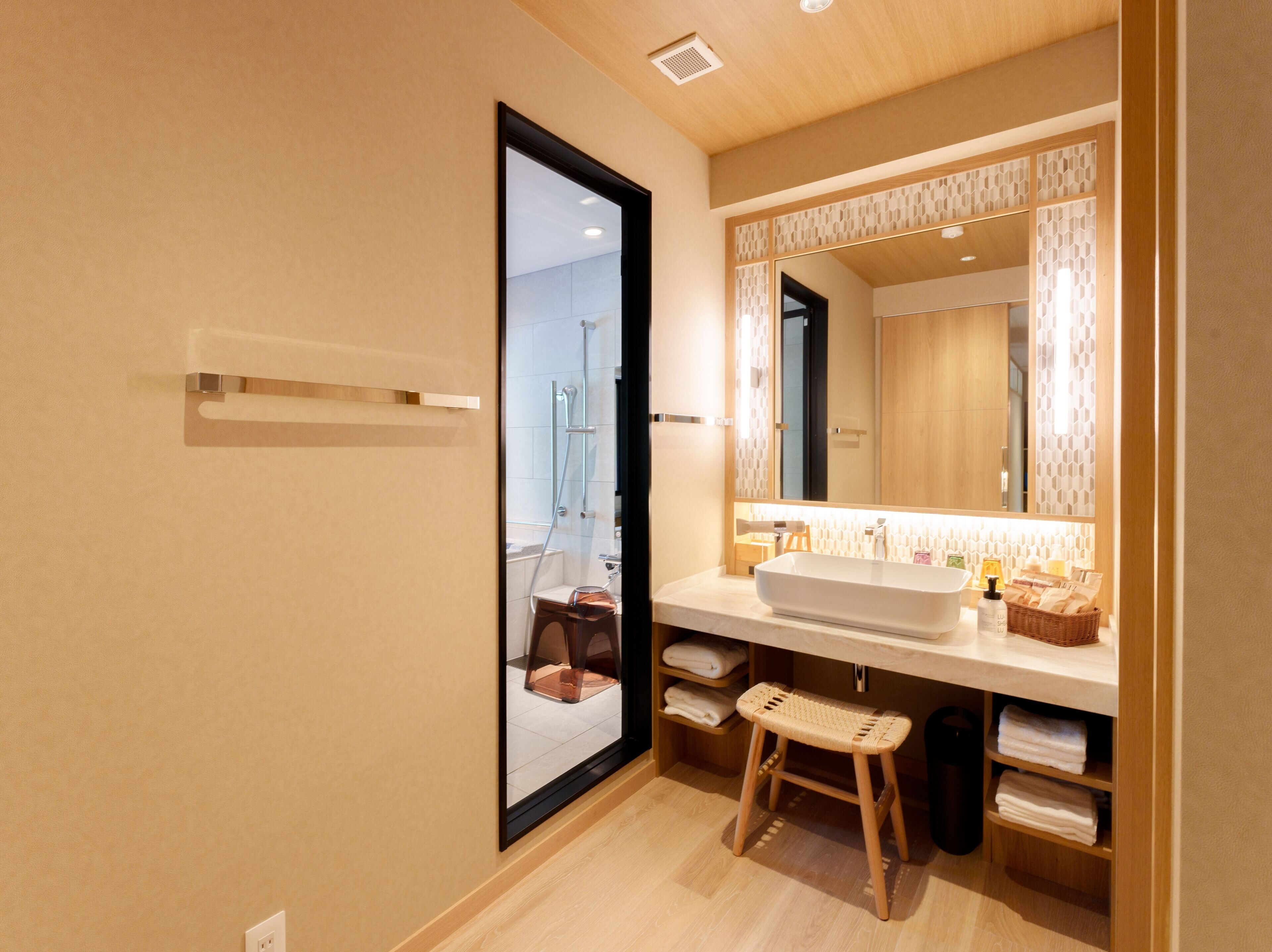 A modern bathroom with a large mirror, a white sink, and a shower. There is a wooden stool and shelves with towels.