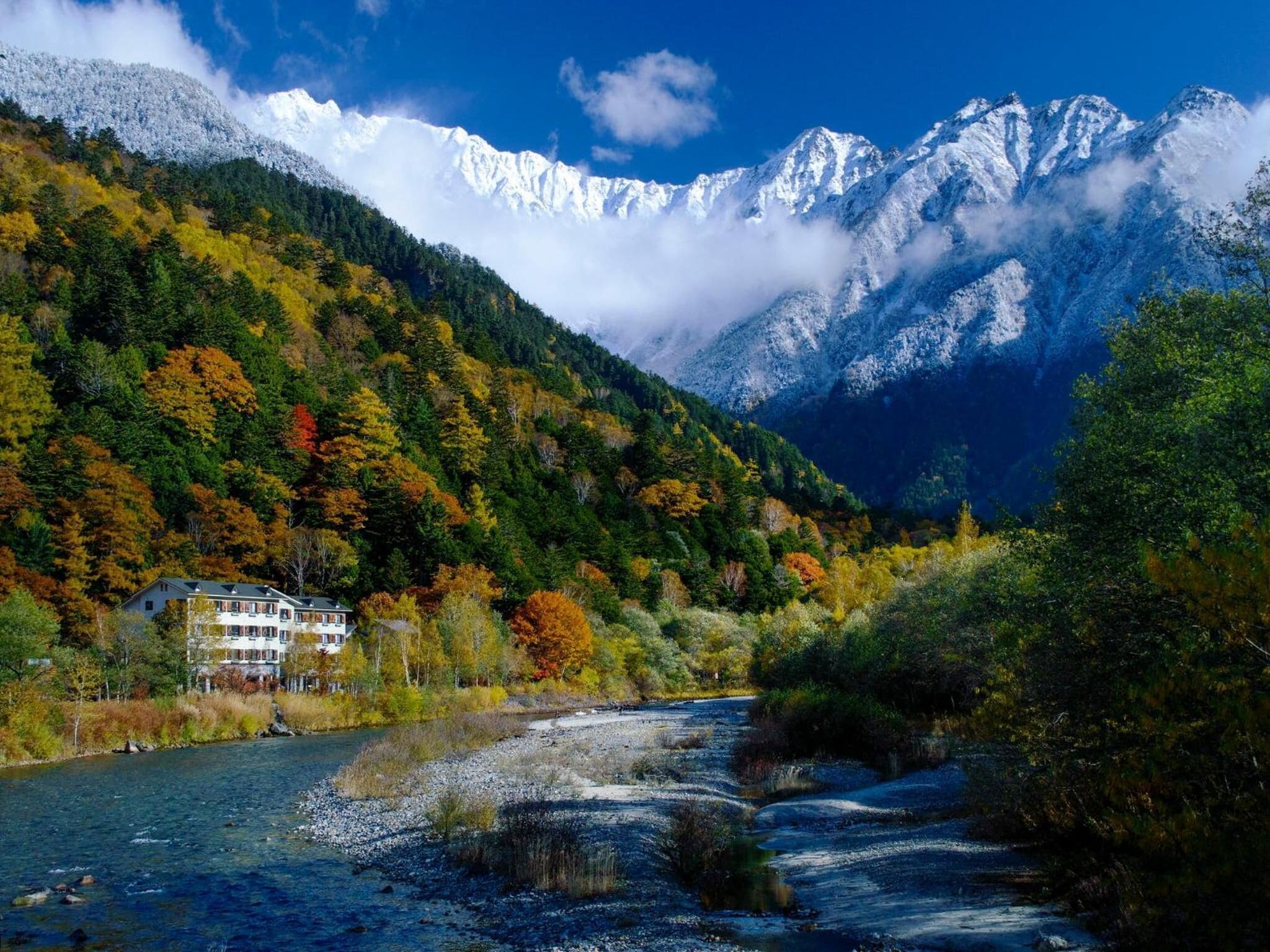 Un paysage de montagne avec une rivière, des arbres aux couleurs automnales et une maison offrant une vue sur les montagnes.