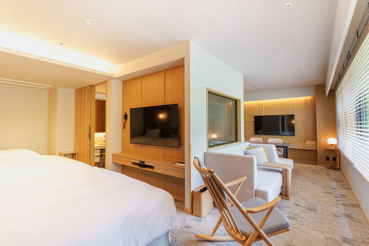 Quarto de hotel moderno com uma cama grande, televisão, área de estar com sofá e poltronas, e um guarda-roupa de madeira.