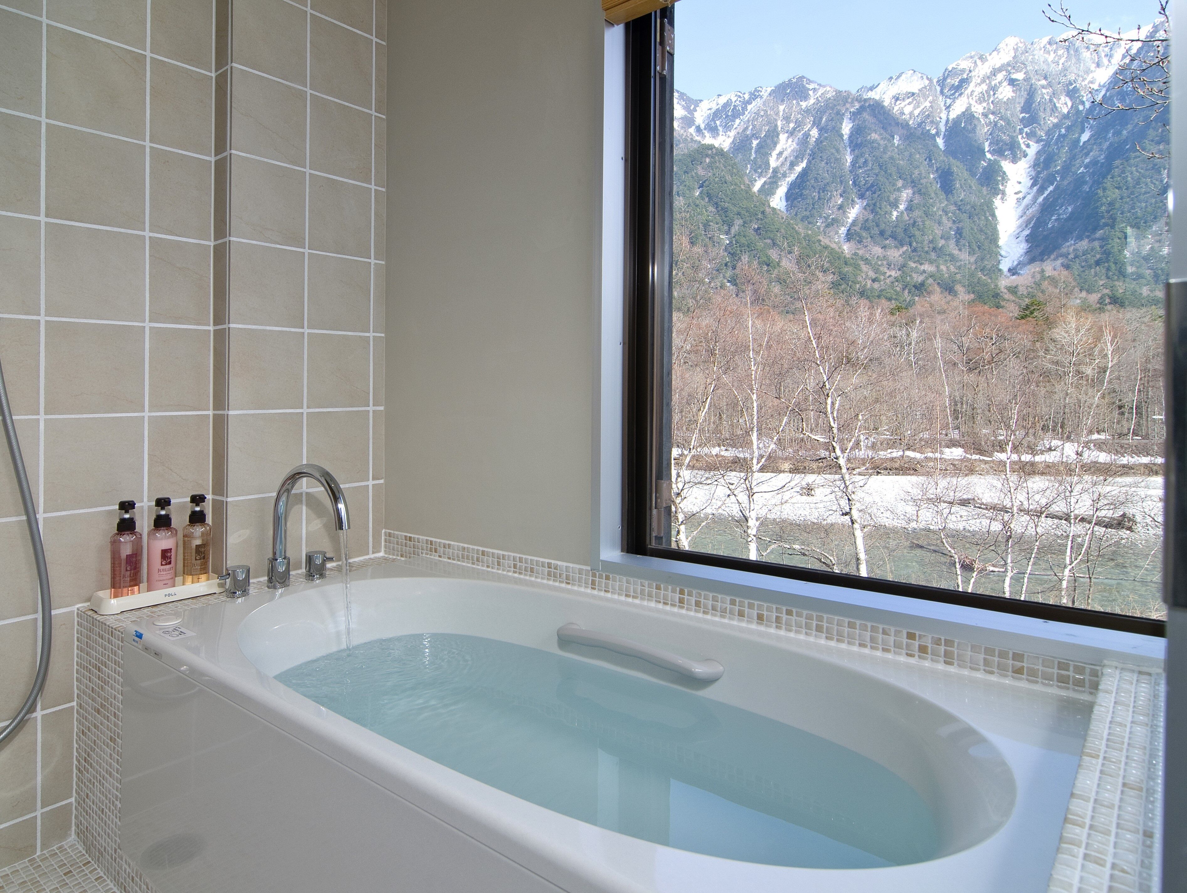 A bathroom with a bathtub, a window with a mountain view, and toiletries on the side.