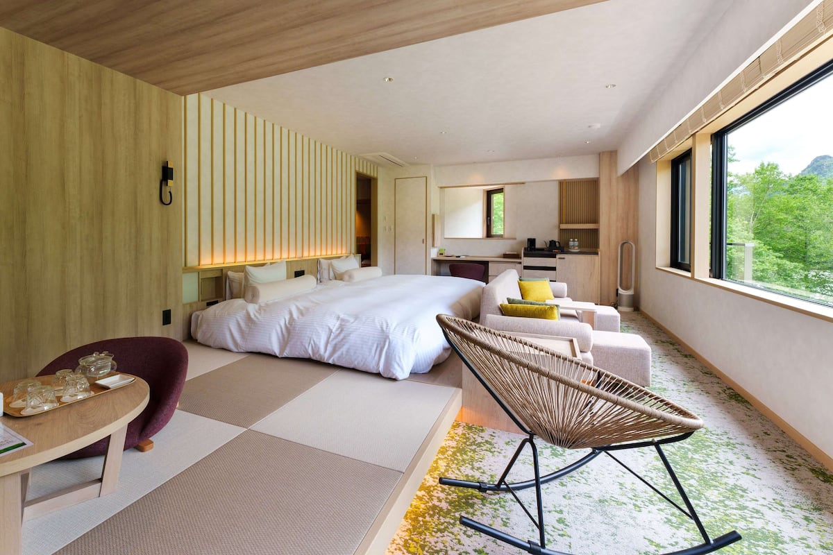 Quarto de hotel moderno com uma cama grande, área de estar com uma poltrona, uma mesinha e vista para o verde lá fora.