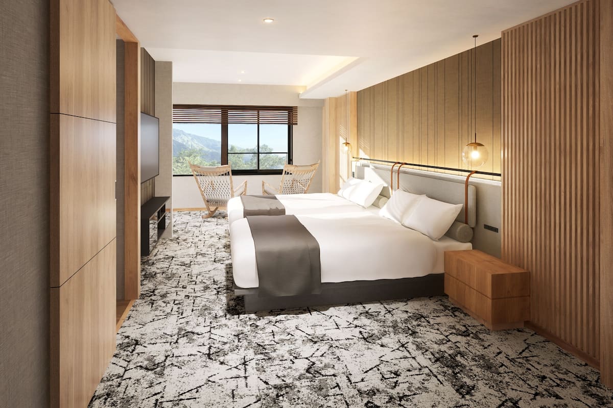 Quarto de hotel moderno com cama grande, duas cadeiras, uma escrivaninha e vista para as montanhas pela janela.