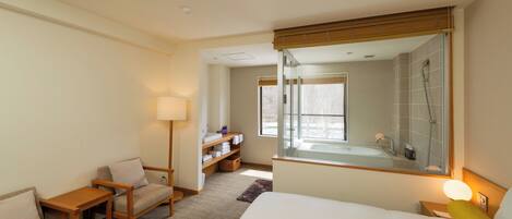 A hotel room with a bed, a desk, a chair, a lamp, a bathroom with a shower, and a large mirror.
