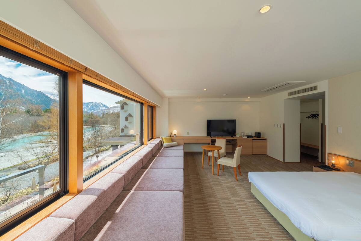 Quarto de hotel moderno com uma janela grande, uma cama, uma escrivaninha com cadeira e uma televisão.