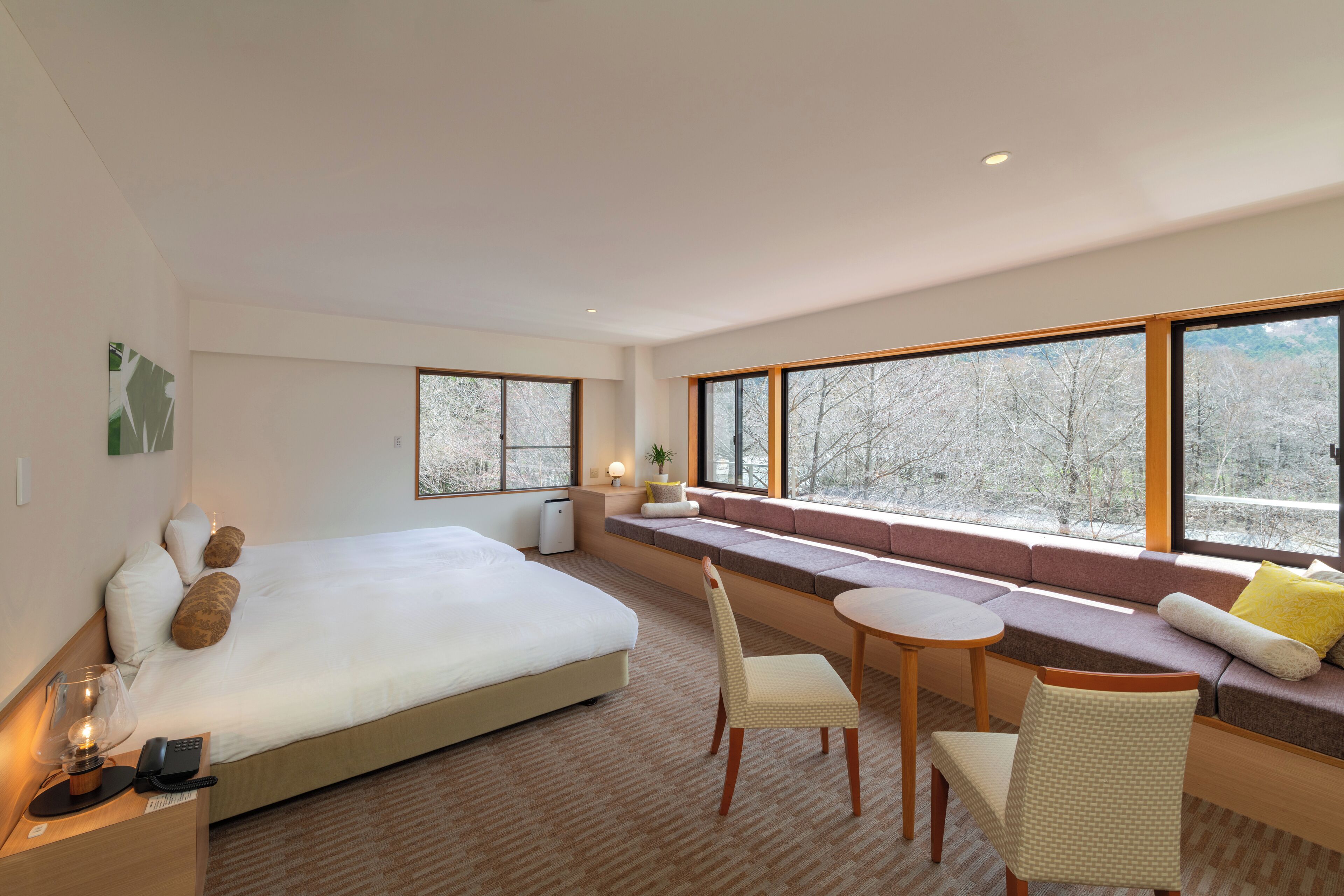 Habitación de hotel moderna con una cama grande, un espacio de descanso con mesa y grandes ventanales con vistas a árboles.