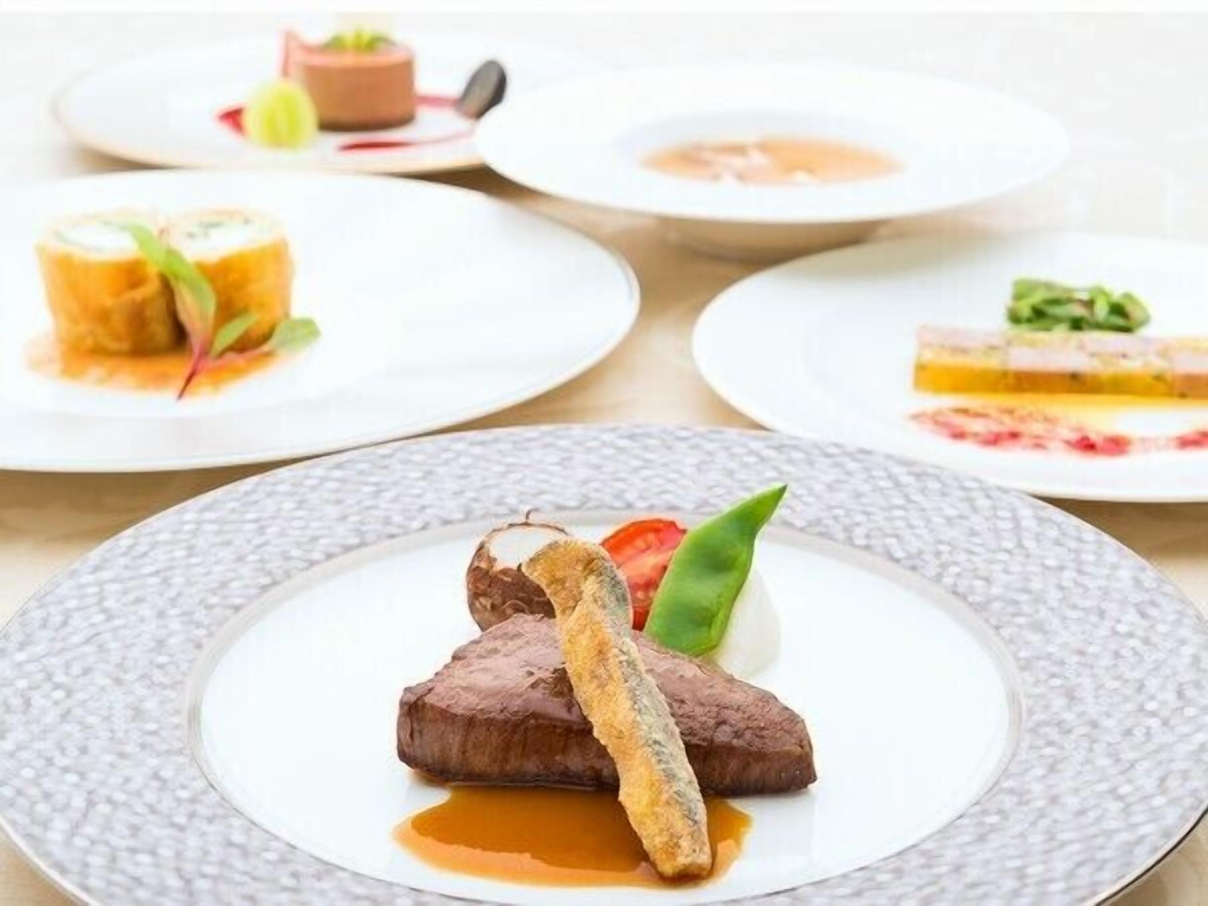 Uma mesa com vários pratos de comida, incluindo um bife, um filé de peixe e molho, além de outros pratos ao fundo.