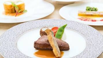 Uma mesa com vários pratos de comida, incluindo um bife, um filé de peixe e molho, além de outros pratos ao fundo.