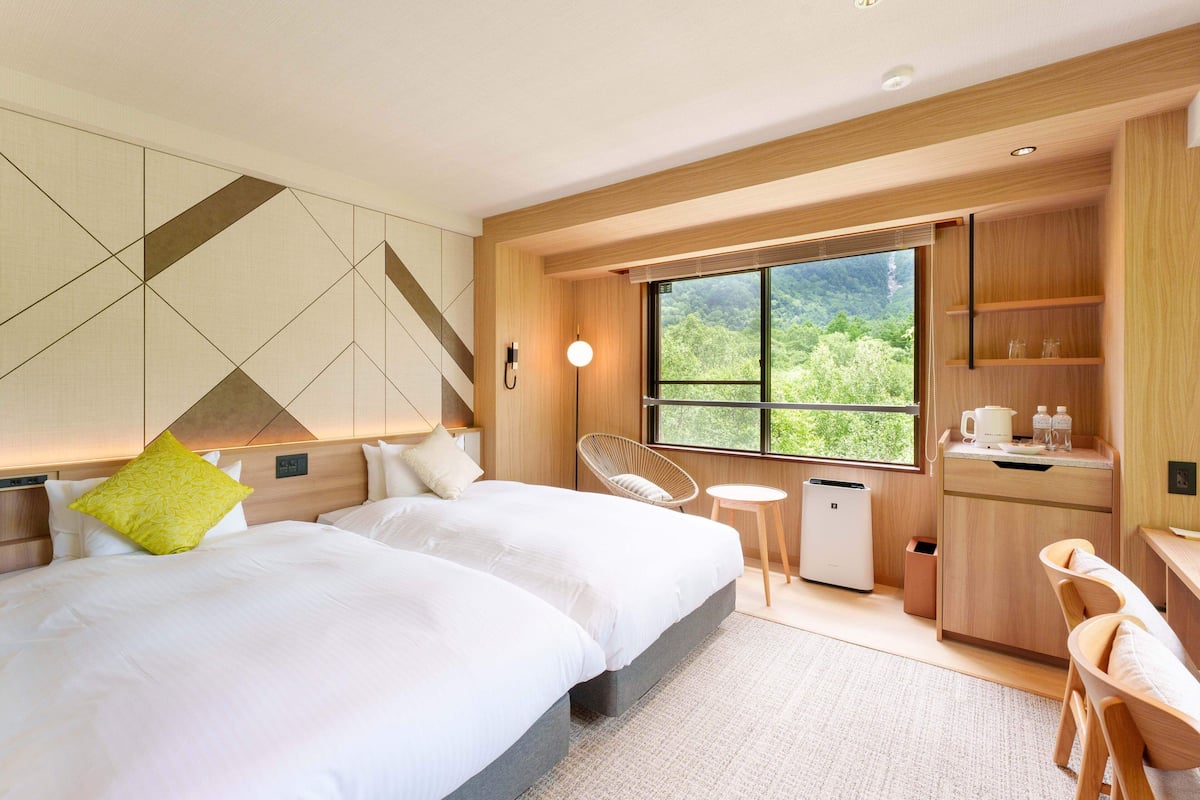 Quarto de hotel moderno com uma cama grande, uma secretária pequena, uma cadeira e uma janela com vista para o verde.