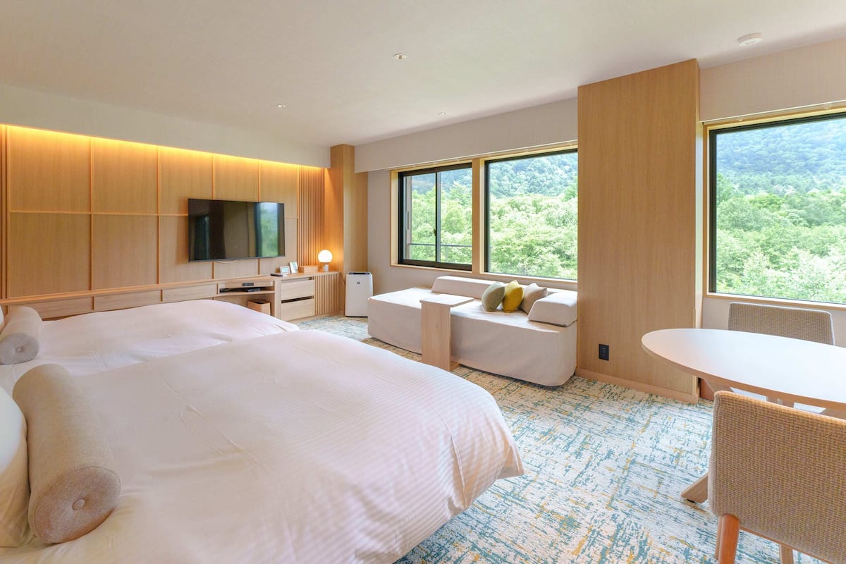 Quarto de hotel moderno com uma cama grande, uma escrivaninha, uma cadeira e uma janela com vista para a floresta.