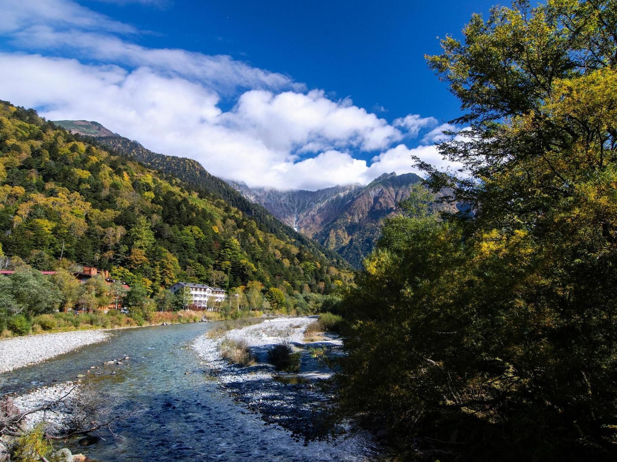 A river flowing through a valley with mountains in the background and a building with multiple windows on the left.