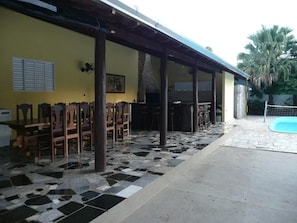 A covered outdoor area with a tiled floor, wooden chairs, a pool, and a yellow wall with a picture.
