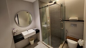 A modern bathroom with a glass-enclosed shower, a round mirror, a white sink, and a toilet. - Cemerlang (Bandung)