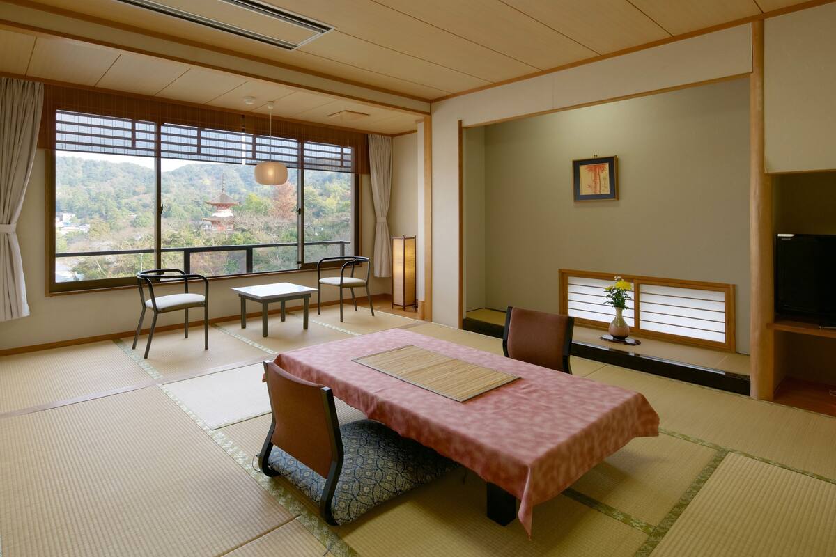 A traditional Japanese room with tatami flooring, a tatami mat table, chairs, a television, and a view of a temple through large windows.