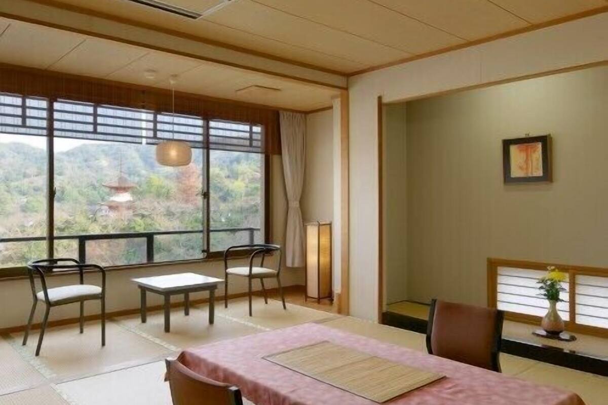 A traditional Japanese-style room with tatami flooring, a low table with a pink tablecloth, and a view of a mountainous landscape through large windows.