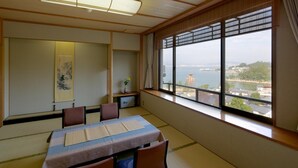 A traditional Japanese room with tatami flooring, a low table with cushions, a shoji screen, and a view of a body of water and buildings. - Jukeiso (Hatsukaichi)