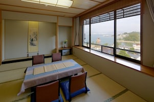 Una habitación japonesa tradicional con suelo de tatami, una mesa baja con cojines, un shoji y vistas a un cuerpo de agua y edificios.