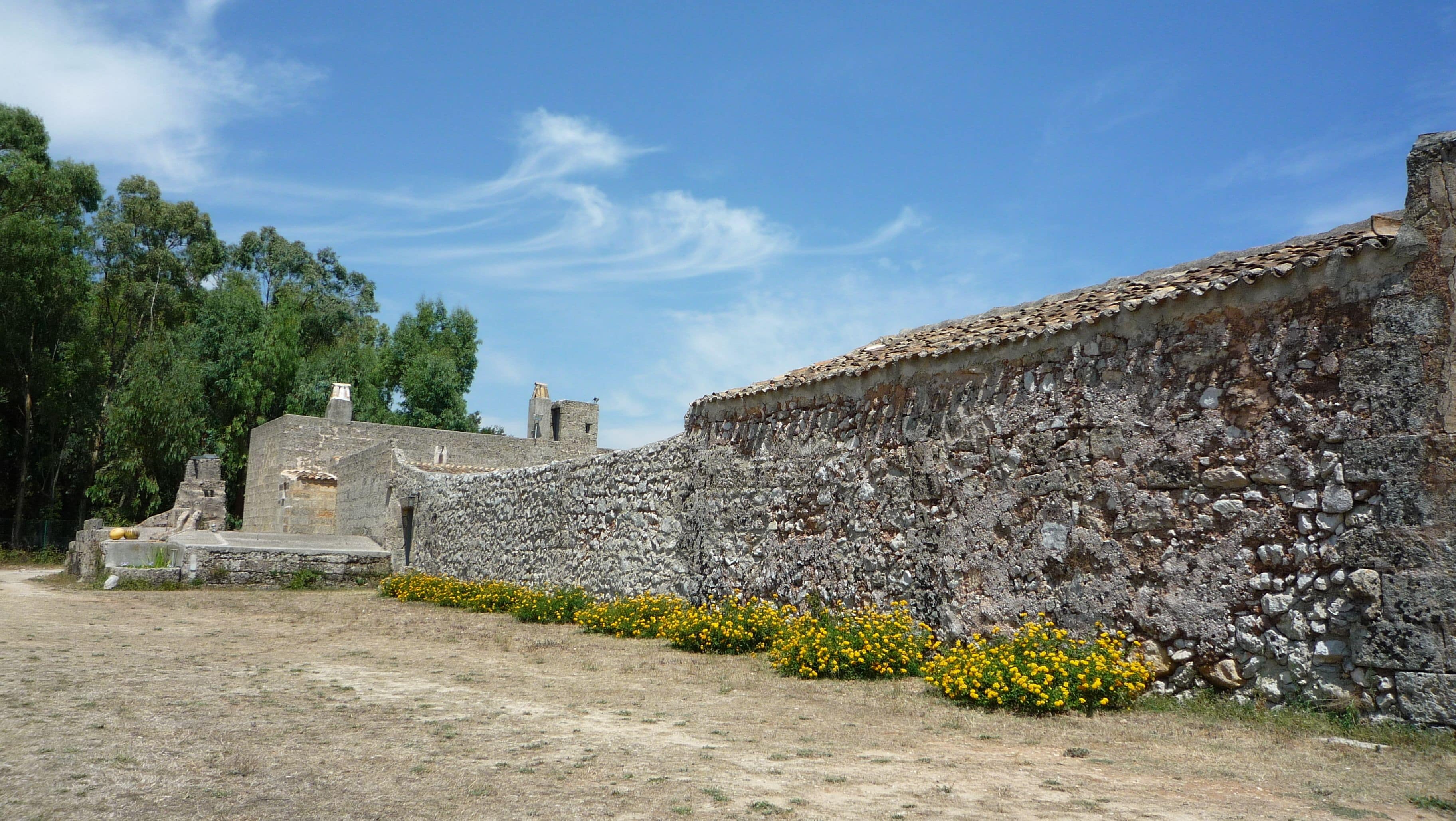 Una antigua pared de piedra con un edificio pequeño y flores amarillas al frente.